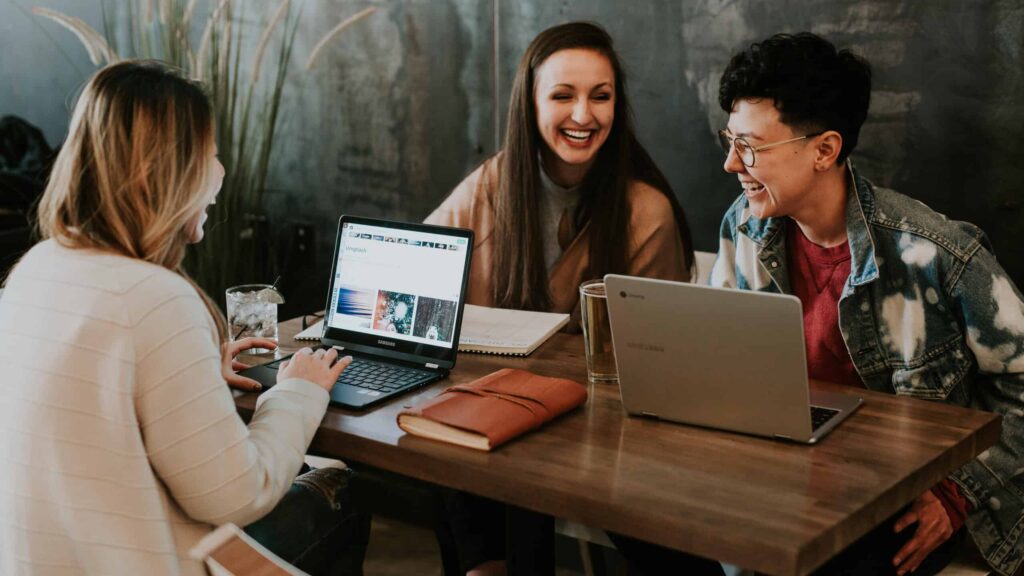 Três pessoas estão sentadas em uma mesa de madeira em um café, sorrindo e trabalhando em laptops. Notebooks e um copo de água também estão na mesa, refletindo uma atmosfera de colaboração e employer branding que promove um ambiente de trabalho alegre.