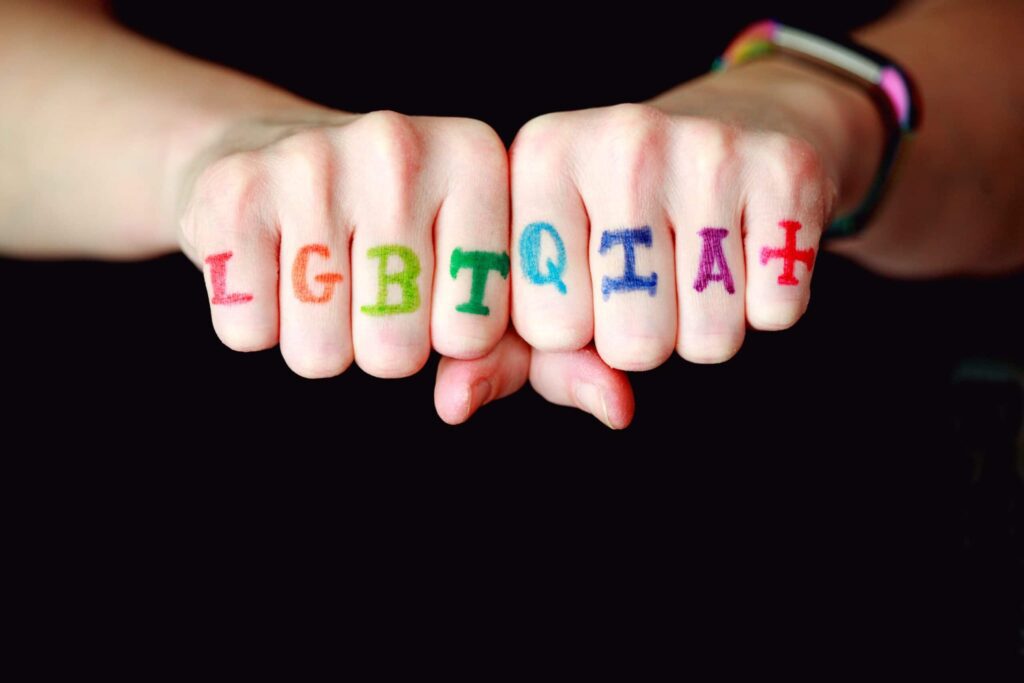 Dois punhos são mostrados com cada letra LGBTQIA+ escrita nos nós dos dedos em cores diferentes. O fundo é preto, destacando as letras coloridas.