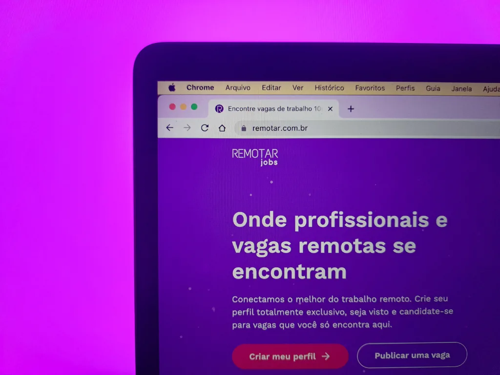 Uma tela de computador exibe um site com o título Remotar jobs e texto em português sobre conectar profissionais com oportunidades de trabalho remoto. A tela está contra um fundo roxo.