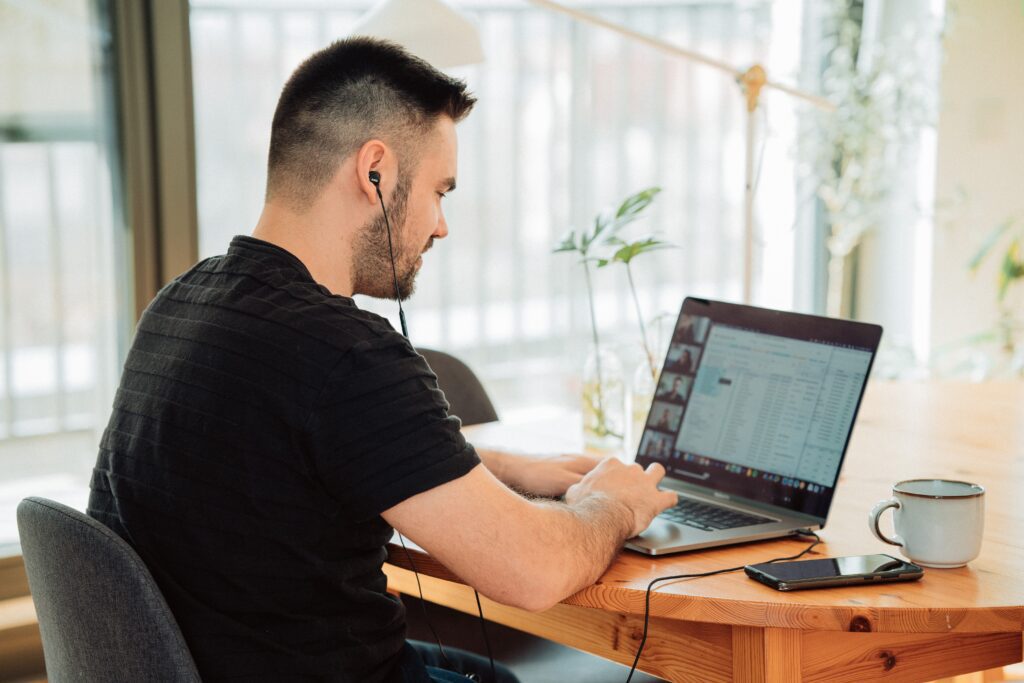 Um homem usando fones de ouvido está sentado em uma mesa de madeira, focado na tela de seu laptop exibindo uma planilha. Um smartphone e uma caneca estão sobre a mesa. A luz natural entra por uma janela próxima.