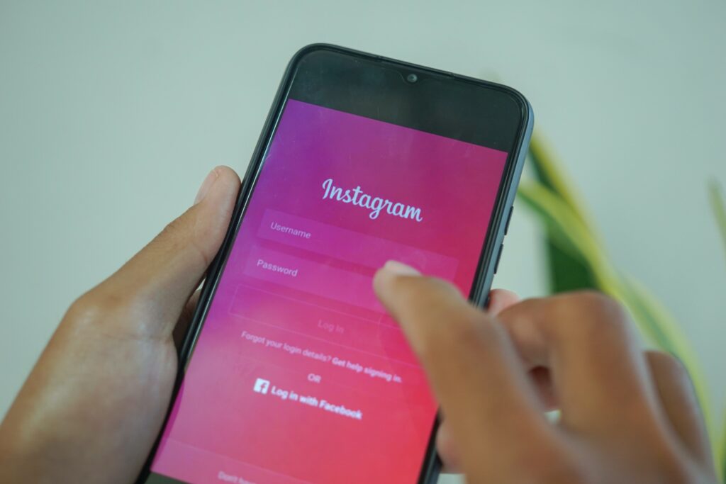 Uma pessoa segurando um smartphone exibindo a tela de login do Instagram. A tela mostra campos para nome de usuário e senha, com opções para fazer login ou usar o Facebook. O dedo da pessoa está pronto para interagir com a tela.