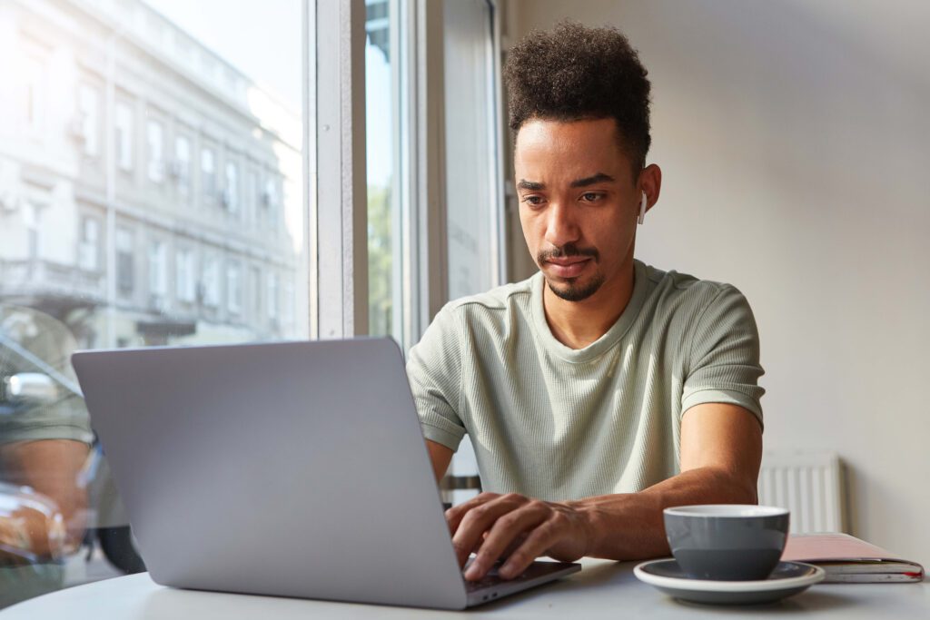 Uma pessoa com cabelo cacheado usando uma camisa verde-claro está sentada em uma mesa em um café, trabalhando em um laptop. Uma xícara de café e um notebook estão sobre a mesa. Grandes janelas oferecem uma vista da rua lá fora.