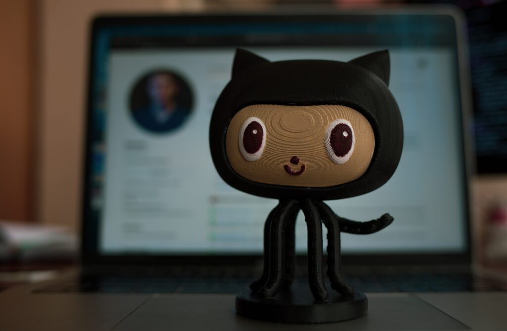 Uma pequena estatueta preta com aparência de gato está em um teclado de laptop. A tela do laptop está desfocada no fundo, mostrando uma página da web com uma foto de perfil e texto.
