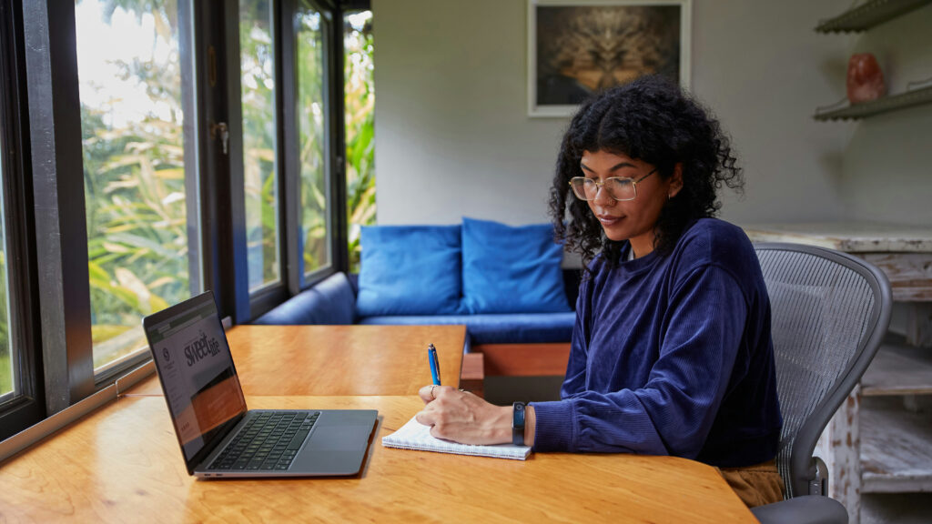 Uma pessoa com cabelo cacheado e óculos está sentada em uma mesa de madeira, escrevendo em um caderno. Eles estão usando um laptop, com janelas mostrando vegetação lá fora. A sala tem um sofá azul e uma foto emoldurada na parede.