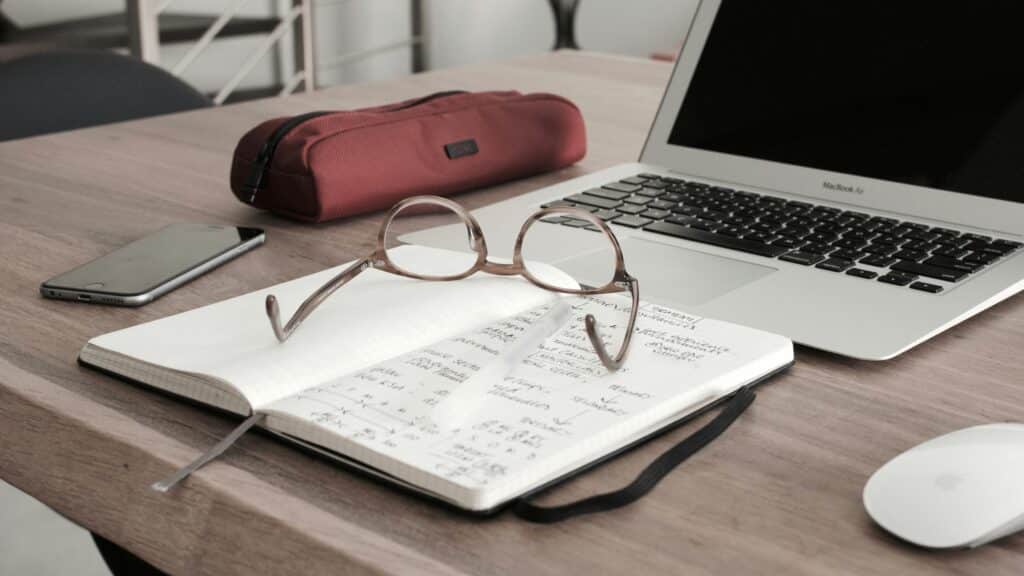 Um par de óculos descansando sobre um caderno aberto com notas manuscritas, lembrando um currículo para o trabalho remoto, são colocados sobre uma mesa de madeira. Perto estão um smartphone, um estojo vermelho, um laptop e um mouse sem fio.