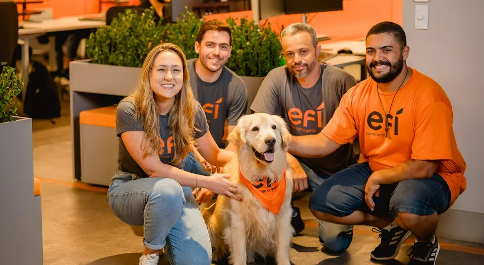 Um grupo de quatro pessoas, três homens e uma mulher, estão sorrindo ao redor de um golden retriever em um ambiente de home office. Eles estão vestindo camisas combinando com um logotipo contra o pano de fundo de plantas verdes e detalhes em laranja, incorporando o espírito de efí em seu espaço moderno e colaborativo.