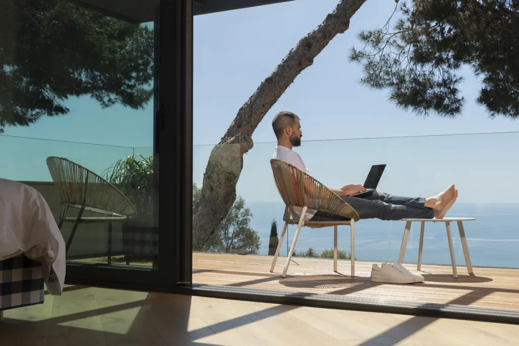 Uma pessoa está sentada em uma cadeira de convés com um laptop, abraçando a vida de vaga internacional e remota enquanto encara uma vista panorâmica do oceano. O convés é adjacente a uma porta de vidro que reflete os móveis internos. Uma árvore se inclina sobre o convés, e o céu está limpo.