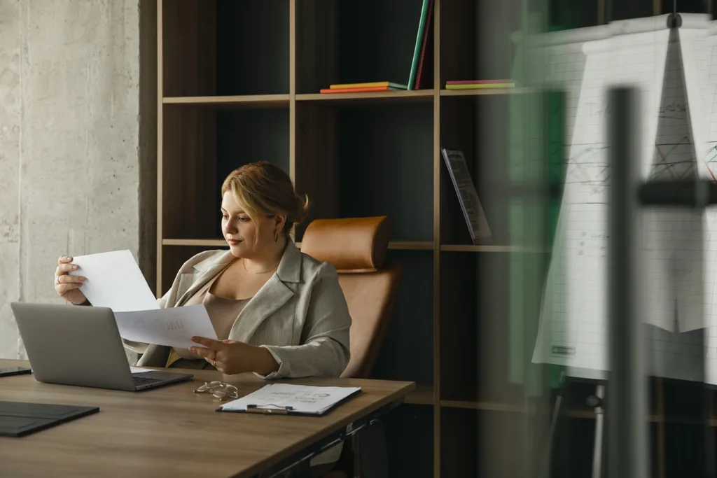Uma pessoa está sentada em uma mesa em seu escritório em casa, examinando documentos com um laptop aberto. Atrás dela, um gráfico em um flip board complementa a prateleira cheia de livros e fichários coloridos. A iluminação é suave e natural, criando um ambiente ideal para tarefas administrativas e financeiras.