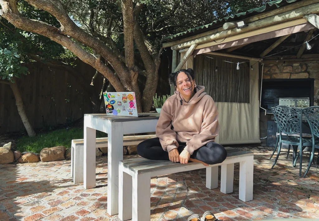 Uma pessoa com um moletom bege está sentada de pernas cruzadas em um banco branco ao ar livre, sorrindo. Abraçando geoarbitragem e trabalho remoto, eles aproveitam o ambiente tranquilo com um laptop coberto de adesivos na mesa atrás deles. A área é sombreada por árvores, com uma estrutura rústica e churrasqueira ao fundo.