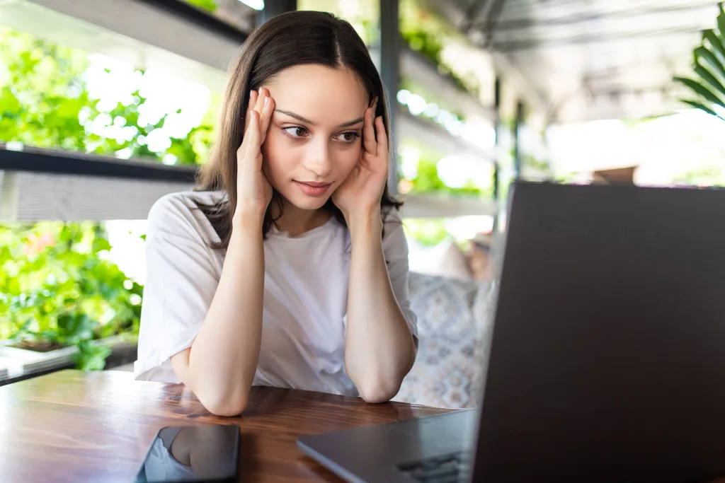 Uma mulher de camisa branca está sentada em uma mesa de madeira, focada em seu laptop enquanto analisa 8 erros comuns ao se candidatar para uma vaga remota. Suas mãos repousam sobre as têmporas, um smartphone está ocioso ao seu lado. Através de grandes janelas, a vegetação exuberante fornece um pano de fundo sereno para sua concentração intensa.