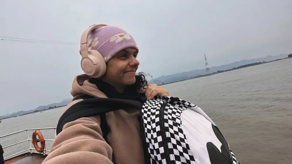 Uma pessoa vestindo um casaco bege, gorro roxo e fones de ouvido sorri enquanto segura uma bolsa xadrez. Eles estão em um barco com um corpo de água e montanhas enevoadas ao fundo sob um céu nublado.