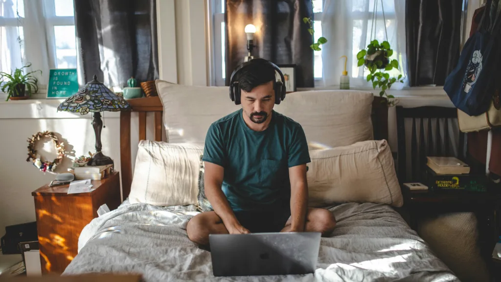 Um homem está sentado de pernas cruzadas em uma cama, fones de ouvido, mergulhando em Cinco mitos e verdades sobre o trabalho remoto em seu laptop. O quarto exala uma atmosfera aconchegante com plantas em vasos, uma luminária decorativa e cortinas emoldurando a janela.