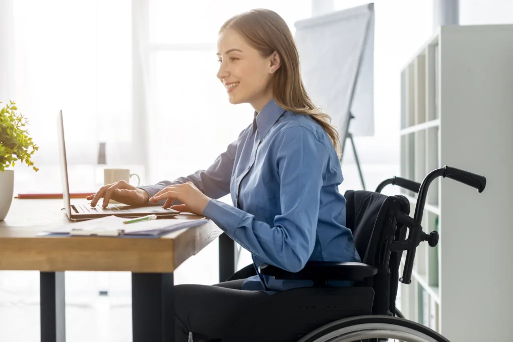 Uma mulher em uma cadeira de rodas trabalha remotamente em uma mesa com seu laptop, sorrindo em uma camisa azul. O fundo apresenta prateleiras e um flip chart, enquanto uma pequena planta adiciona calor ao seu espaço de trabalho. Trabalho remoto melhora a vida de pessoas com deficiência?.