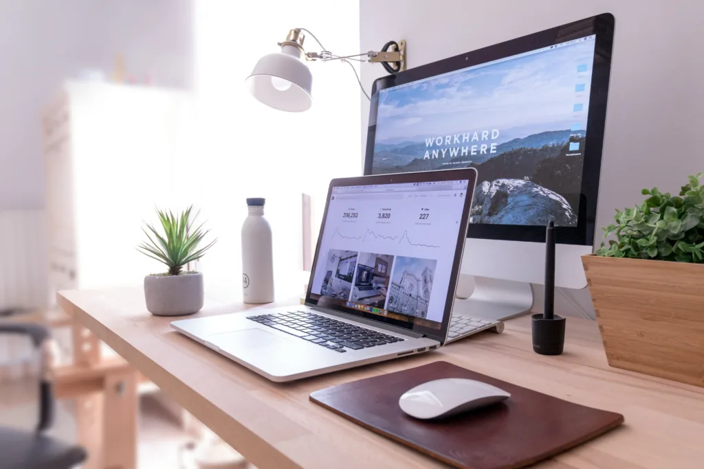 Um espaço de trabalho organizado exibe um laptop aberto em uma página de análise ao lado de uma tela de desktop exibindo "Work Hard Anywhere" e apresentando um compilado de vagas remotas. A mesa também ostenta uma pequena planta, garrafa de água, mouse em um pad, mesa digitalizadora e luminária de mesa — perfeito para oportunidades de homeoffice.