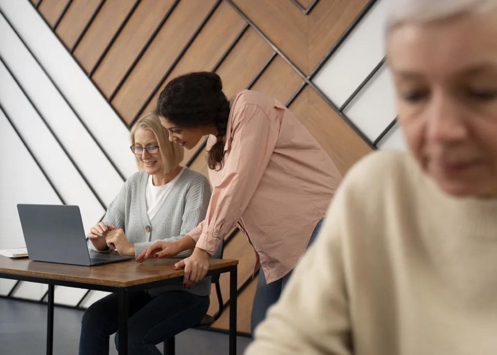Duas mulheres estão colaborando em uma mesa com um laptop. Uma digita enquanto a outra fica de pé, apontando para a tela, talvez discutindo como explicar o trabalho remoto para sua mãe. Uma figura borrada está sentada perto no ambiente de escritório moderno.