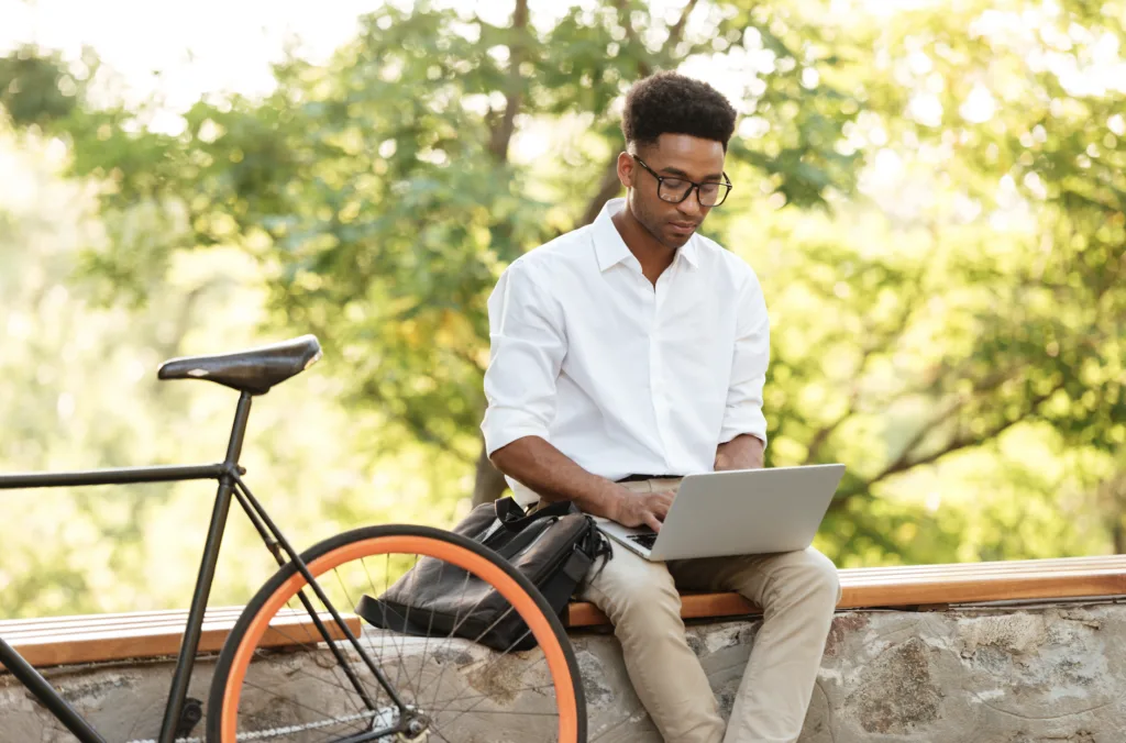 Um homem de camisa branca e óculos está sentado em um muro de pedra, abraçando trabalho remoto enquanto usa um laptop. Perto dali, uma bicicleta com rodas laranja descansa calmamente. O fundo é preenchido com árvores verdes e frondosas se aquecendo sob a luz do céu ensolarado, despertando prazeres adormecidos.