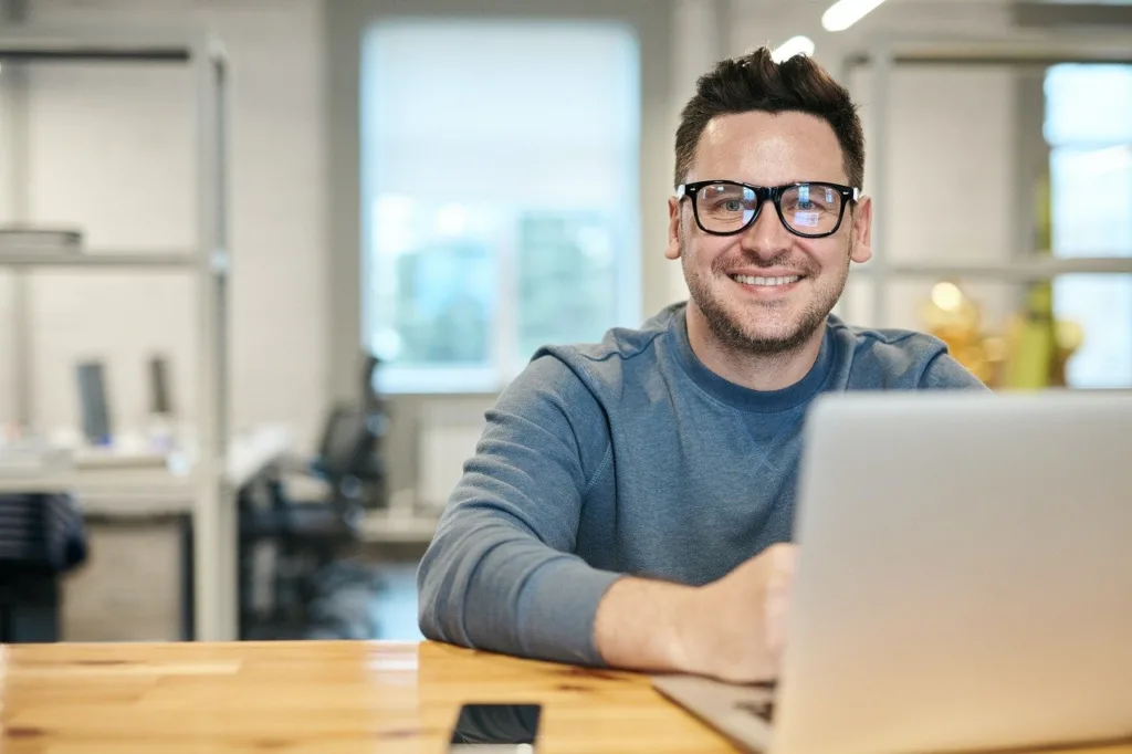 Um homem usando óculos e um suéter azul está sentado em uma mesa de madeira com um laptop aberto. Ele está sorrindo, e há um smartphone por perto. O fundo é um ambiente de escritório com mesas e cadeiras.