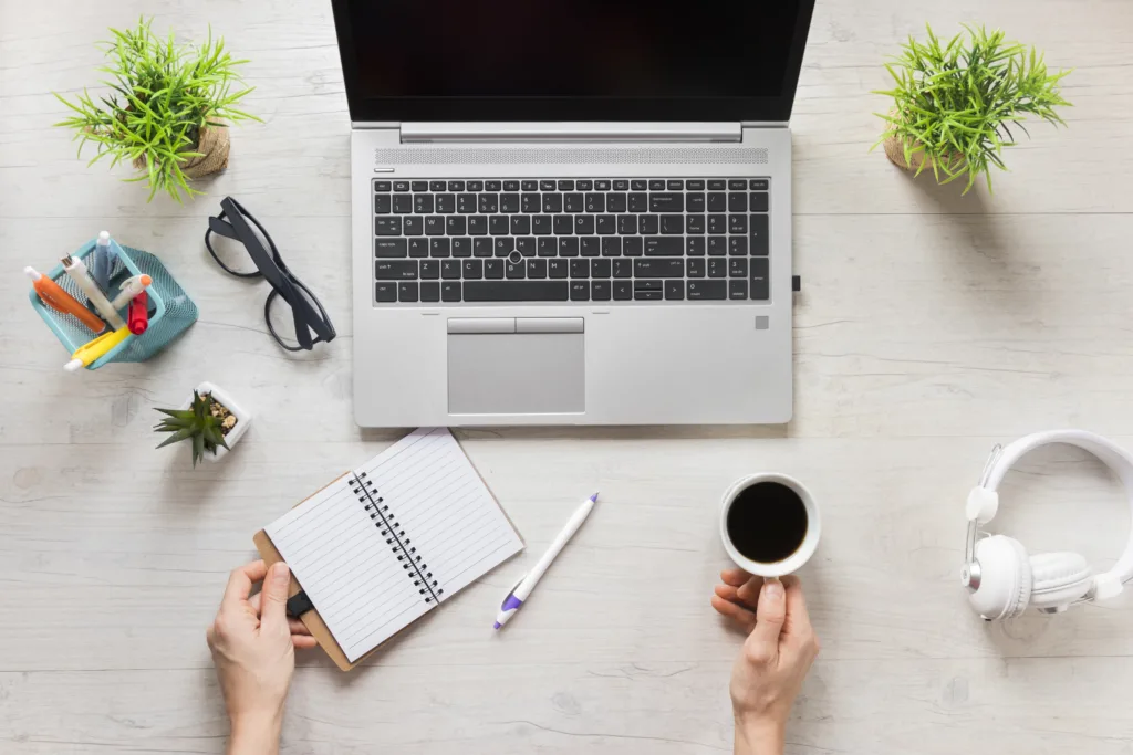 Uma vista superior de um espaço de trabalho com um laptop, óculos escuros, plantas em vasos, um caderno sobre vagas remotas em Marketing e Vendas, uma caneta, café e fones de ouvido brancos em uma mesa de madeira clara. Duas mãos são visíveis; uma segura o caderno enquanto a outra embala o café.