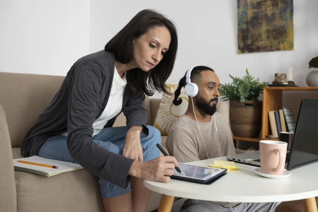 Em uma sala de estar aconchegante repleta de plantas, uma mulher de cardigã cinza e jeans trabalha em seu tablet na mesa de centro, incorporando a essência do trabalho remoto em 2025. Ao lado dela, um homem com fones de ouvido digita em seu laptop, com uma caneca de café por perto.