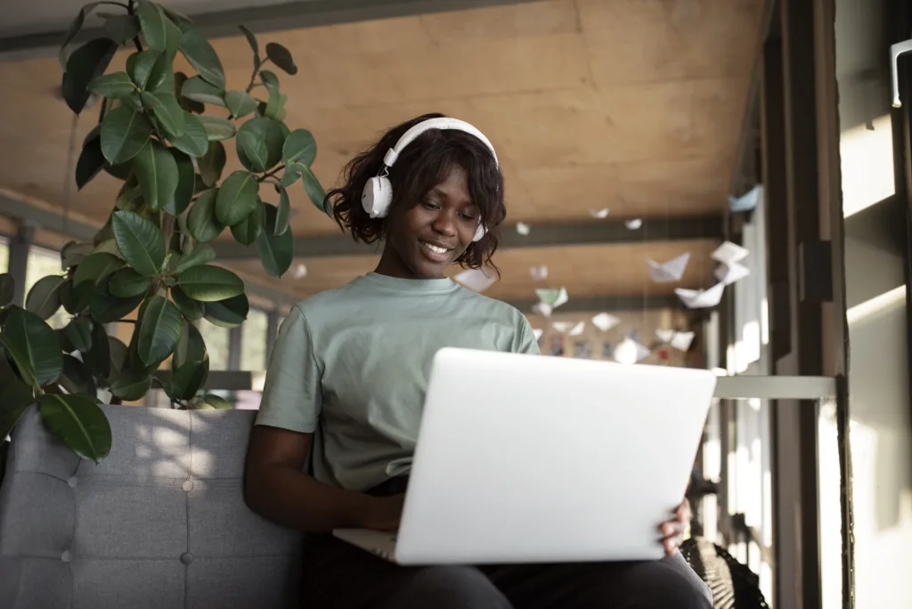 Uma mulher de camisa verde e fones de ouvido está sentada em um sofá cinza, usando seu laptop para explorar vagas remotas em marketing. Ela está sorrindo e focada em meio ao cenário sereno com uma grande planta em vaso e tsurus de papel pendurados no teto em seu quarto bem iluminado.