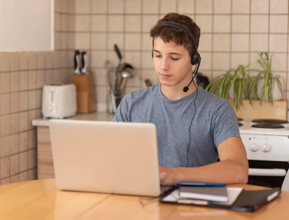 Jovem está sentado em uma mesa, trabalhando em frente a um laptop branco, com fone de ouvido e microfone. Foto para ilustrar um artigo sobre vagas remotas em atendimento e suporte.