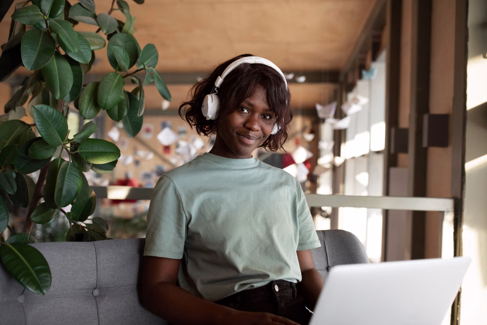 Uma jovem usando fones de ouvido está sentada em um sofá cinza, sorrindo para a câmera, com um laptop aberto à sua frente. Ela está em um ambiente interno, perto de uma planta grande e de um ambiente de trabalho moderno e iluminado.
