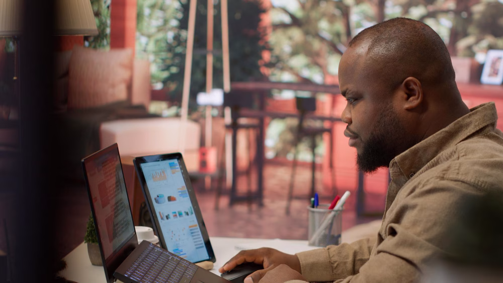 Um homem senta-se em uma mesa trabalhando em um laptop com um segundo monitor que exibe gráficos coloridos. O plano de fundo mostra um espaço interno aconchegante com plantas e móveis.
