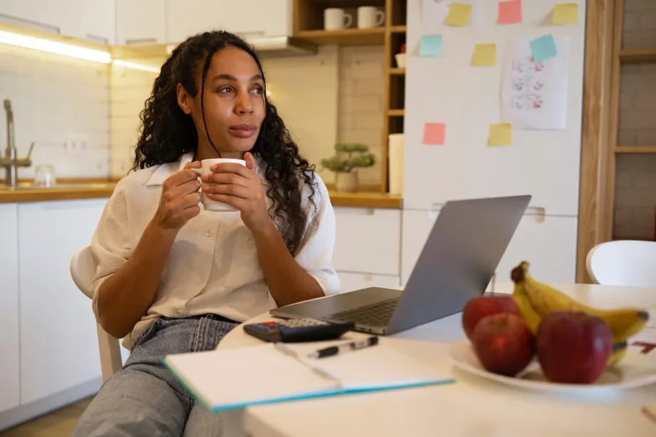 Uma pessoa com cabelos longos e cacheados está sentada à mesa da cozinha, segurando uma caneca e olhando para o lado, ponderando vagas remotas júnior. Um laptop, caderno, caneta e um prato de frutas estão sobre a mesa. A cozinha tem armários brancos e uma geladeira adornada com post-its coloridos.
