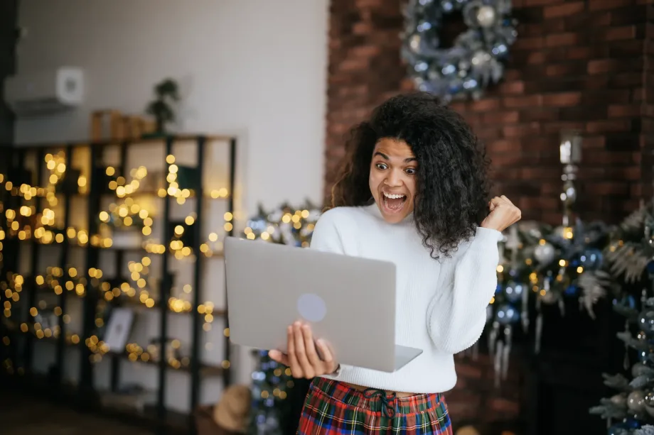 Uma pessoa com cabelo cacheado e um suéter branco olha animadamente para um laptop, personificando a alegria de uma festa de final de ano no trabalho remoto. Eles estão em uma sala de estar decorada de forma aconchegante com luzes festivas e uma guirlanda em uma parede de tijolos, sugerindo uma celebração de temporada de férias.