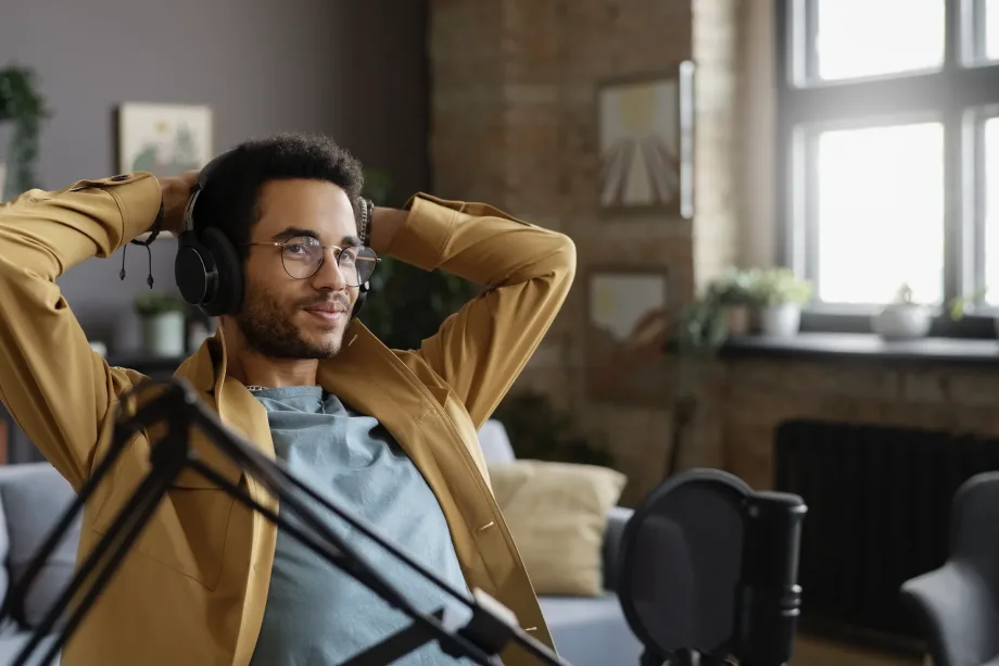 Uma pessoa relaxada, possivelmente gravando podcasts sobre trabalho remoto, senta-se em uma cadeira usando fones de ouvido e uma jaqueta marrom. Na frente deles, há um microfone em um suporte. A sala tem decoração aconchegante e moderna, com plantas e grandes janelas que deixam entrar luz natural.