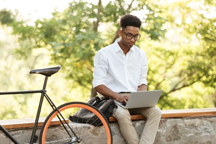 Um homem de camisa branca e óculos está sentado em um muro de pedra, abraçando trabalho remoto enquanto usa um laptop. Perto dali, uma bicicleta com rodas laranja descansa calmamente. O fundo é preenchido com árvores verdes e frondosas se aquecendo sob a luz do céu ensolarado, despertando prazeres adormecidos.