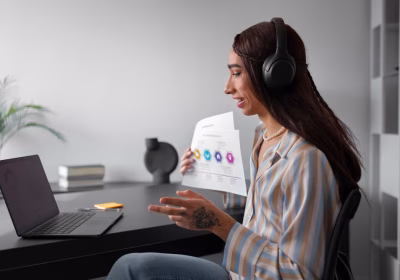 Uma pessoa usando fones de ouvido segura um gráfico impresso enquanto está sentada em uma mesa com um laptop, parecendo participar de uma reunião ou apresentação virtual.