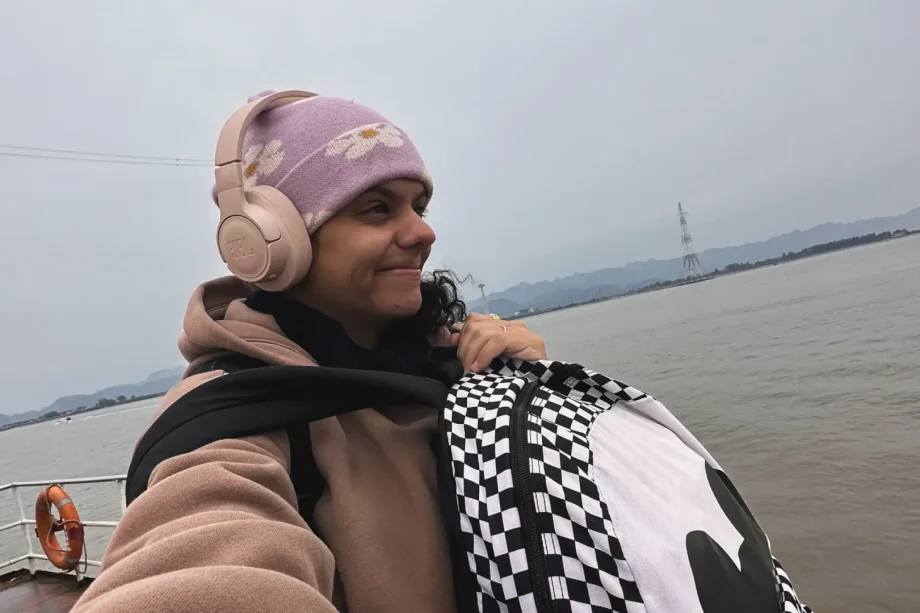 Uma pessoa vestindo um casaco bege, gorro roxo e fones de ouvido sorri enquanto segura uma bolsa xadrez. Eles estão em um barco com um corpo de água e montanhas enevoadas ao fundo sob um céu nublado.