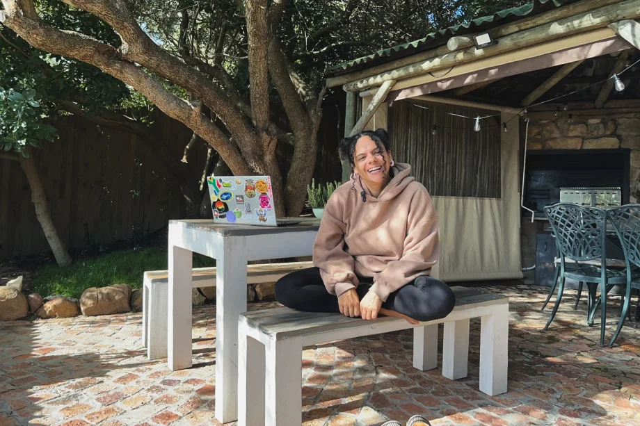 Uma pessoa com um moletom bege está sentada de pernas cruzadas em um banco branco ao ar livre, sorrindo. Abraçando geoarbitragem e trabalho remoto, eles aproveitam o ambiente tranquilo com um laptop coberto de adesivos na mesa atrás deles. A área é sombreada por árvores, com uma estrutura rústica e churrasqueira ao fundo.