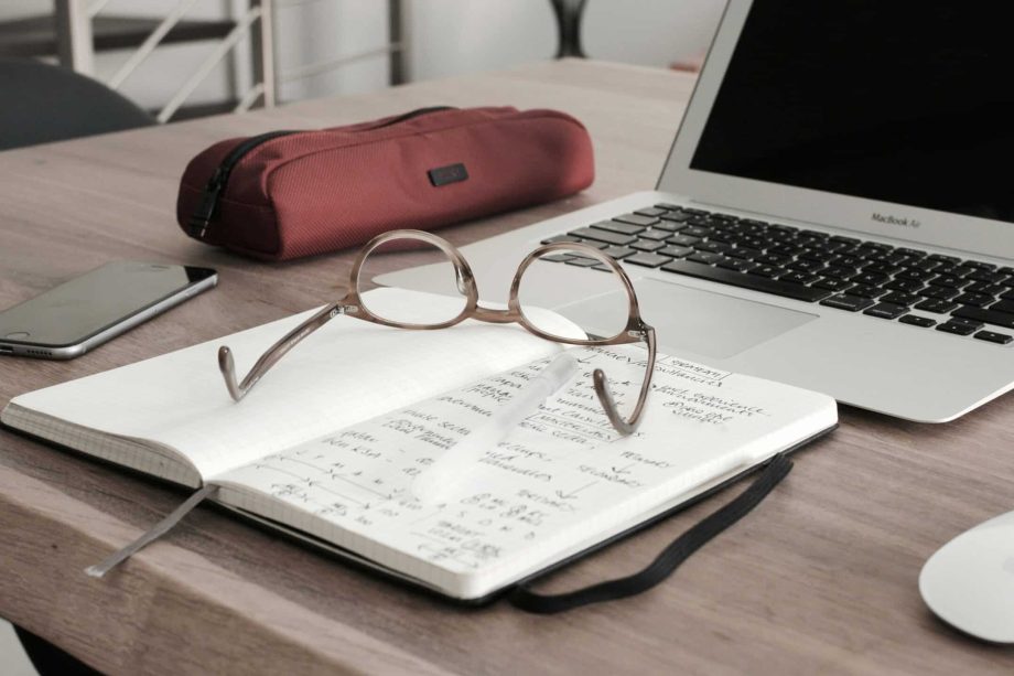 Um par de óculos descansando sobre um caderno aberto com notas manuscritas, lembrando um currículo para o trabalho remoto, são colocados sobre uma mesa de madeira. Perto estão um smartphone, um estojo vermelho, um laptop e um mouse sem fio.