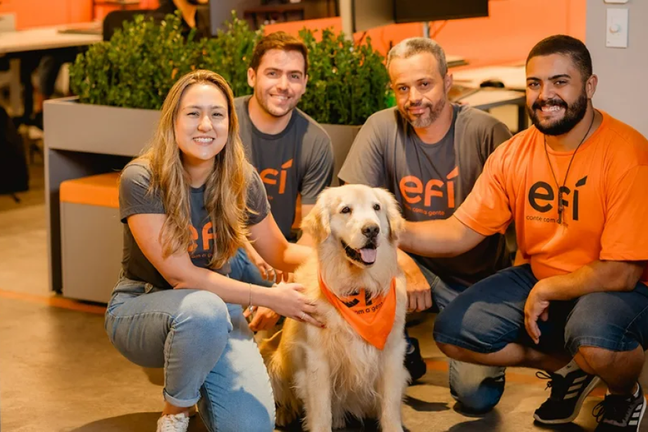 Um grupo de quatro pessoas, três homens e uma mulher, estão sorrindo ao redor de um golden retriever em um ambiente de home office. Eles estão vestindo camisas combinando com um logotipo contra o pano de fundo de plantas verdes e detalhes em laranja, incorporando o espírito de efí em seu espaço moderno e colaborativo.