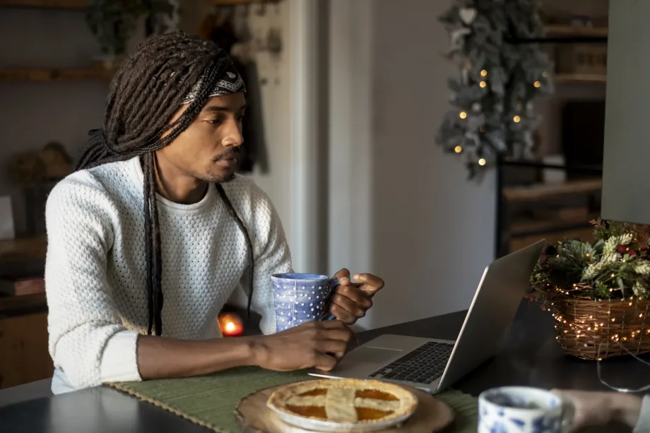 Uma pessoa de cabelos longos está sentada à mesa, segurando uma caneca azul e olhando para um laptop, personificando a essência do trabalho remoto em 2024. Uma torta e uma pequena decoração festiva estão próximas, com luzes desfocadas ao fundo, criando uma atmosfera aconchegante.