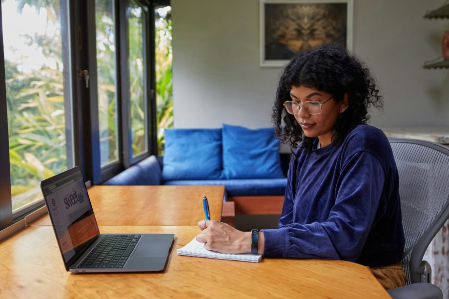 Uma pessoa com cabelo cacheado e óculos está sentada em uma mesa de madeira, escrevendo em um caderno. Eles estão usando um laptop, com janelas mostrando vegetação lá fora. A sala tem um sofá azul e uma foto emoldurada na parede.