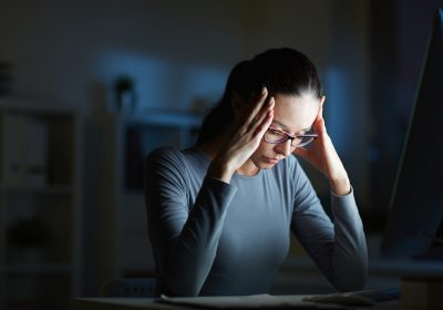 Young businesswoman with headache touching her head while sitting in front of computer monitor by her workplace at night