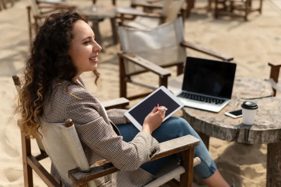 Uma mulher com cabelos cacheados está sentada em uma cadeira de madeira na praia, segurando um tablet e uma caneta. Um laptop, um smartphone e uma xícara de café repousam sobre a mesa rústica à sua frente. Ela sorri, apreciando o ambiente externo enquanto explora países que contratam brasileiros para trabalho remoto.