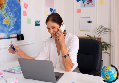 Uma mulher está falando no telefone, à frente de um laptop prateado. No canto esquerdo, temos um mapa colado na parede e uma série de post-its. No canto direito, temos um globo terrestre em miniatura.