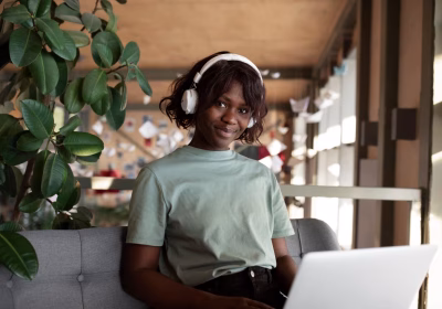 Uma jovem usando fones de ouvido está sentada em um sofá cinza, sorrindo para a câmera, com um laptop aberto à sua frente. Ela está em um ambiente interno, perto de uma planta grande e de um ambiente de trabalho moderno e iluminado.